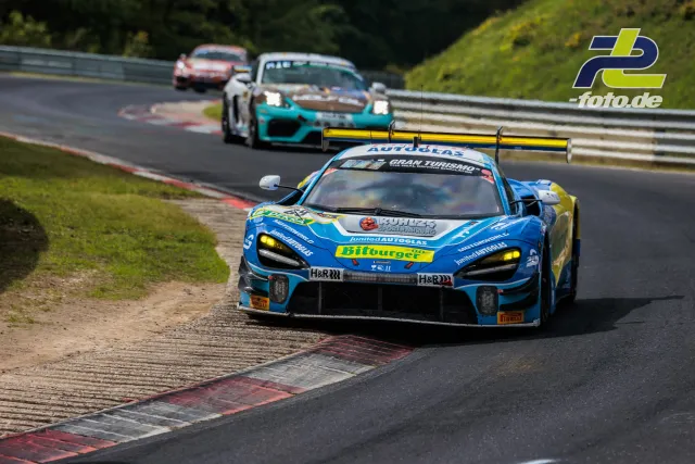 McLaren 720S GT3 in blau-gelber Lackierung fährt über Randsteine auf der Nordschleife, gefolgt von weiteren GT3-Fahrzeugen.