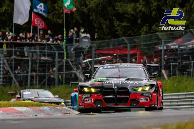 BMW M4 GT3 in schwarz-rot auf der Nordschleife, im Hintergrund Zuschauertribüne und weitere Rennwagen