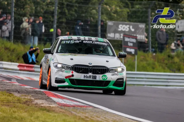 Weißer BMW mit grünen Akzenten und orangen Kerbs auf der Nordschleife beim 24h-Rennen Nürburgring, Zuschauer im Hintergrund.