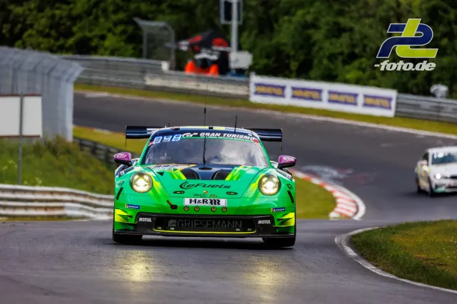 Grün-gelber Porsche 911 GT3 R mit eingeschaltetem Licht auf nasser Nordschleife während des Qualifyings, gefolgt von weiteren Fahrzeugen.