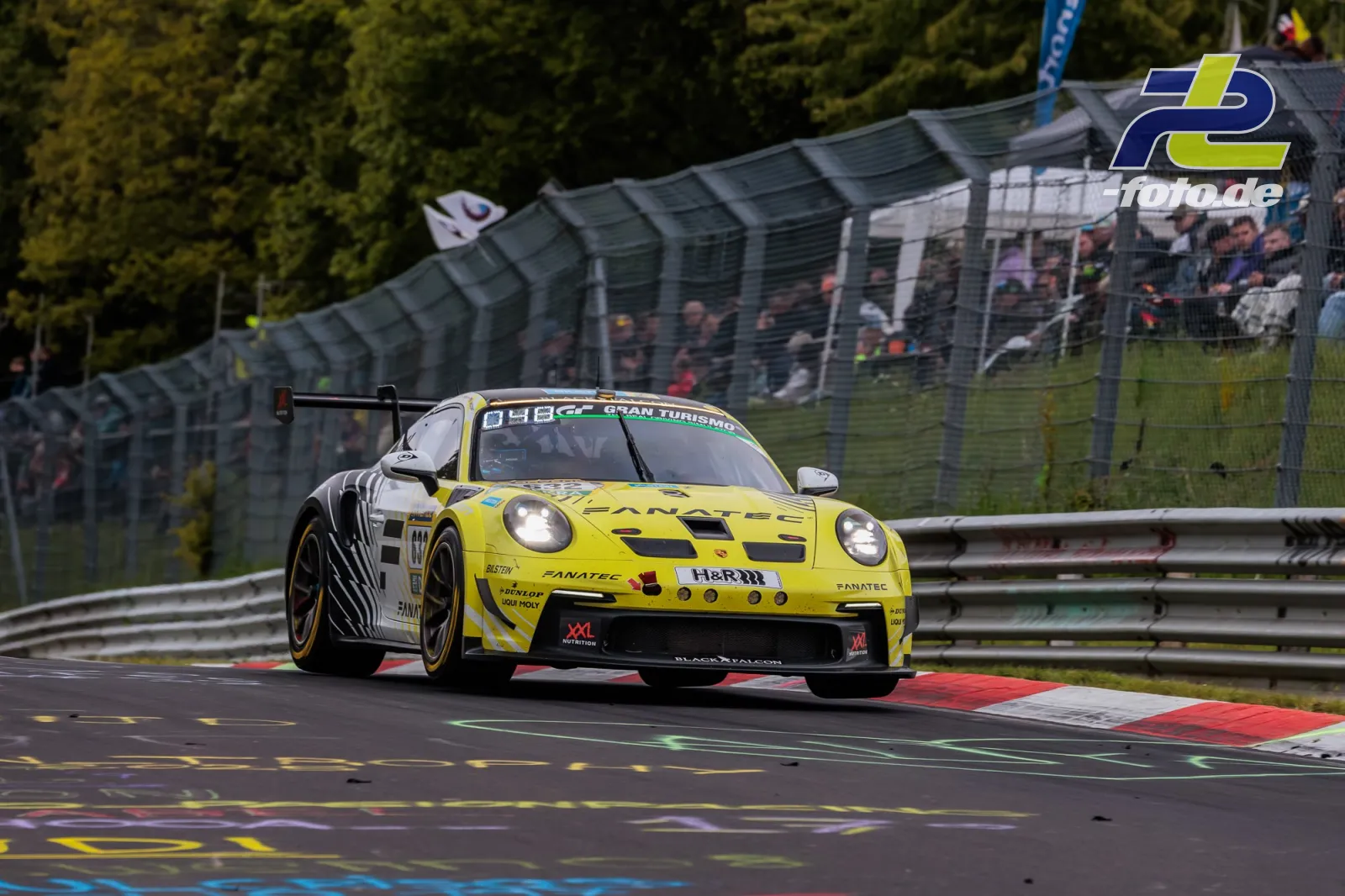 Gelber Porsche GT3 mit Startnummer 632 auf der Nordschleife während des 24h-Rennens, Zuschauertribüne im Hintergrund.