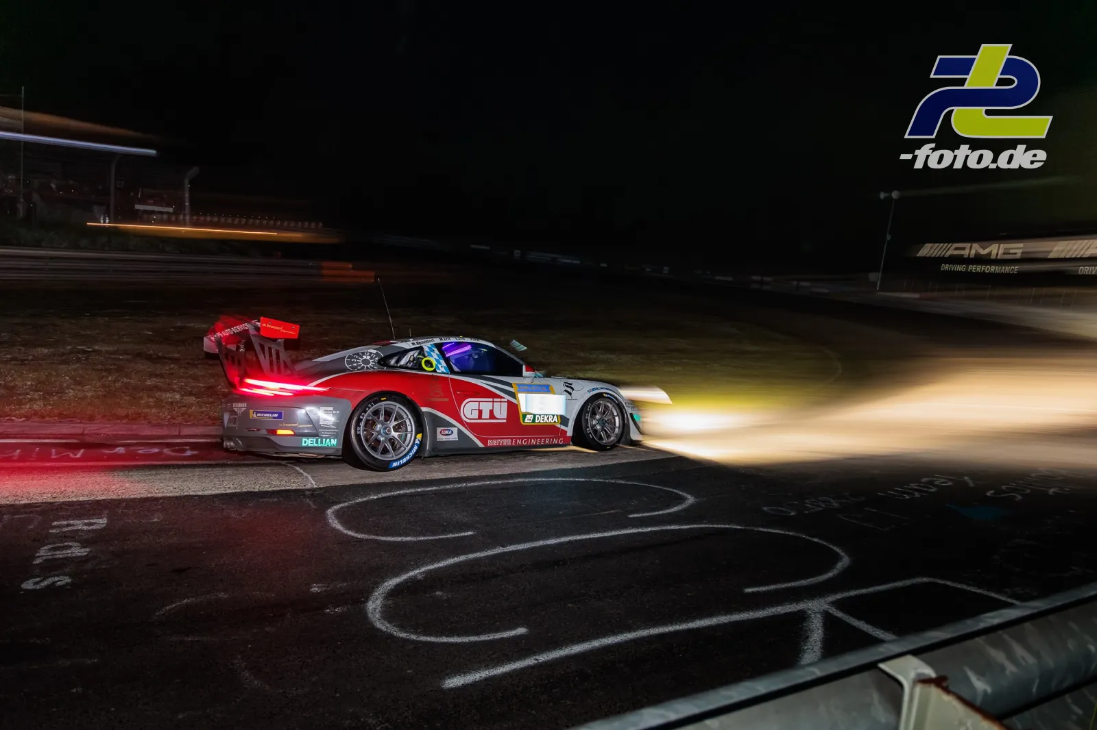 Porsche 911 GT3 Cup bei Nacht auf dem Nürburgring, rot-weiße Lackierung, beleuchtete Scheinwerfer, Kurvenausgang mit Randstein.