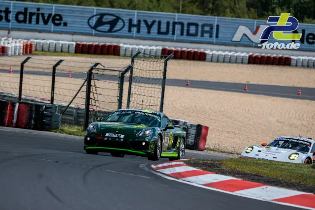 Porsche Cayman GT4 in dunkelgrün mit neongrünen Akzenten auf der Nordschleife, im Hintergrund ein Porsche 911 GT3 im Kiesbett.