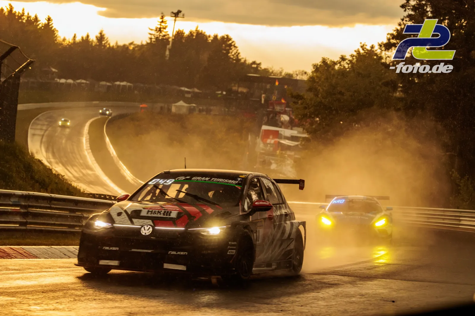 VW Golf auf nasser Nordschleife bei Gegenlicht, weitere Fahrzeuge folgen durch dramatische Gischt bei Sonnenuntergang.