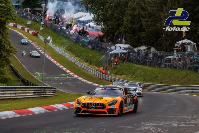Mercedes-AMG GT4 in orange-schwarzer Lackierung auf der Nürburgring-Nordschleife während der Formationsrunde, Zuschauer mit Rauchfackeln im Hintergrund.