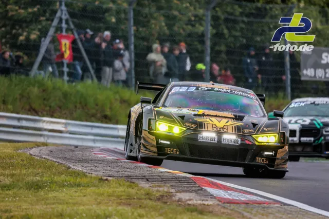 GT3-Rennwagen mit gold-schwarzer Lackierung auf der Nordschleife während des 24h-Rennens, verfolgt von einem weiteren Fahrzeug.