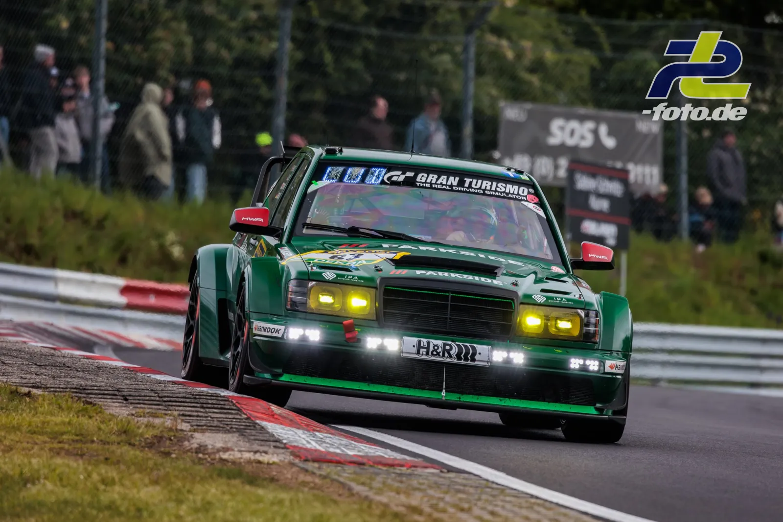 Grüner Mercedes-Benz 190 E mit gelben Scheinwerfern in Kurve auf dem Nürburgring, Zuschauer hinter Zaun im Hintergrund