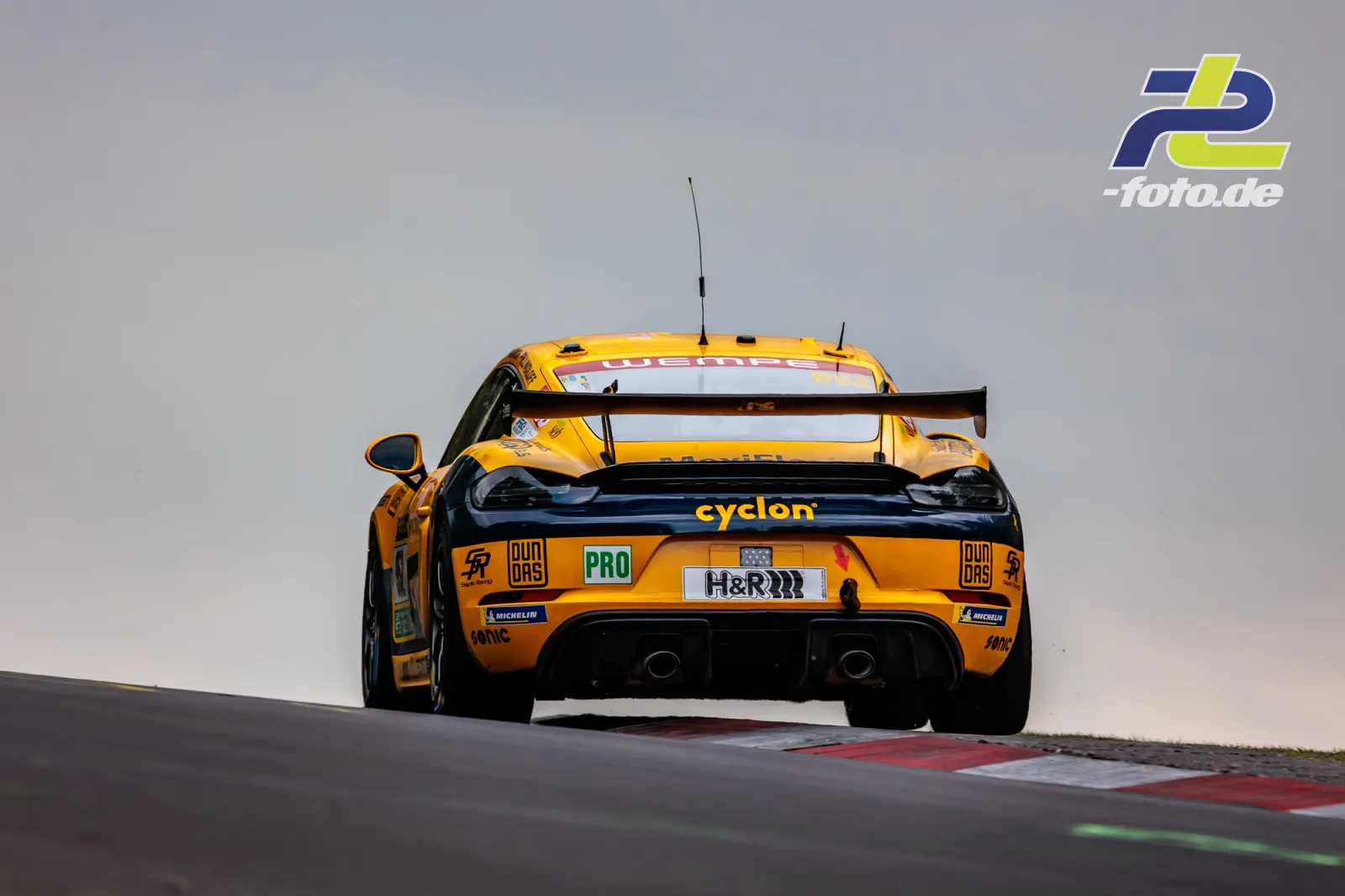 Gelber Porsche Cayman GT4 mit cyclon-Lackierung von hinten an einer Kuppe auf der Nürburgring-Nordschleife beim 24h-Rennen.