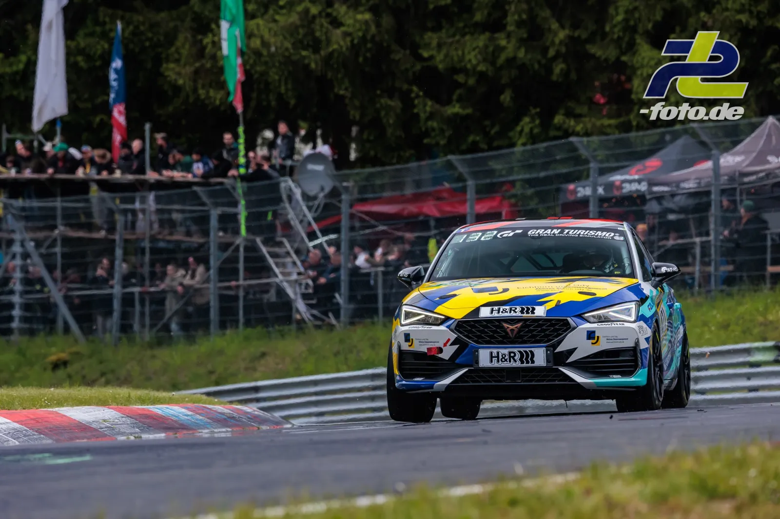 Cupra Leon KL in Kurve beim 24h-Rennen am Nürburgring, Tribüne und Zelten im Hintergrund, gelb-blaue Lackierung