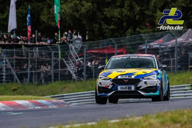 Cupra Leon KL in Kurve beim 24h-Rennen am Nürburgring, Tribüne und Zelten im Hintergrund, gelb-blaue Lackierung