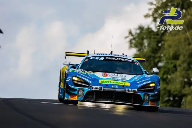 Blauer McLaren 720S GT3 mit gelben Akzenten im Rennen über eine Kuppe auf dem Nürburgring, Tageslicht.