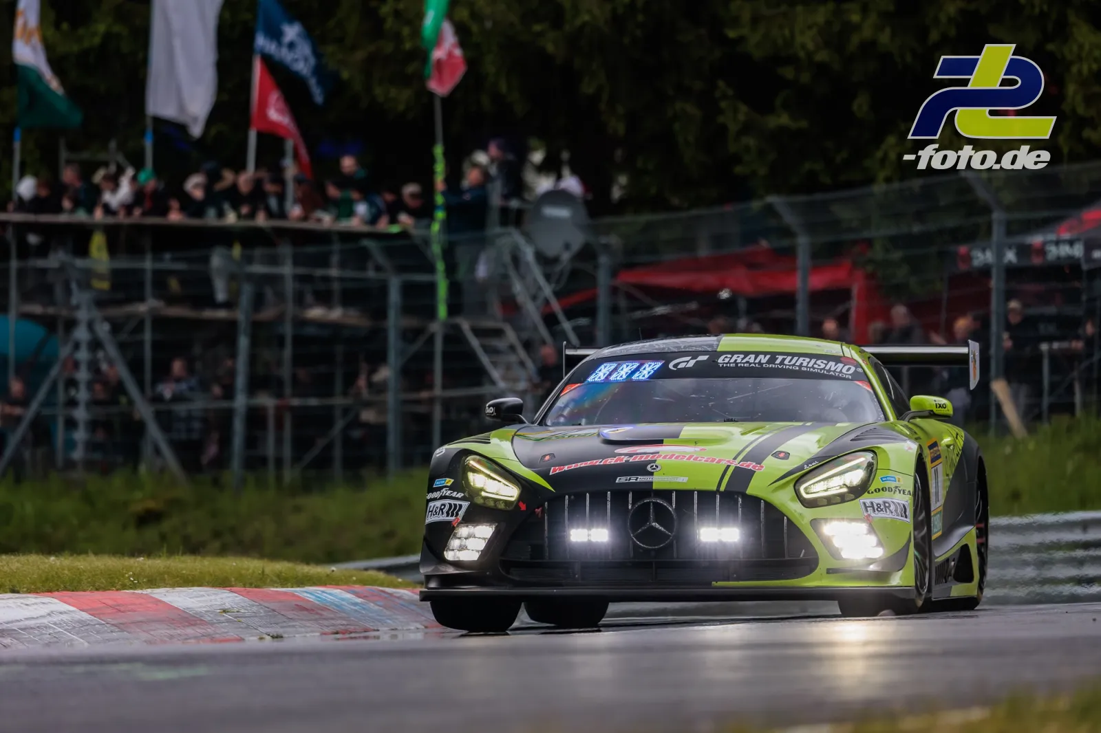 Mercedes-AMG GT3 in neongelber Lackierung mit Licht während des 24h-Rennens am Nürburgring, Zuschauertribüne im Hintergrund.