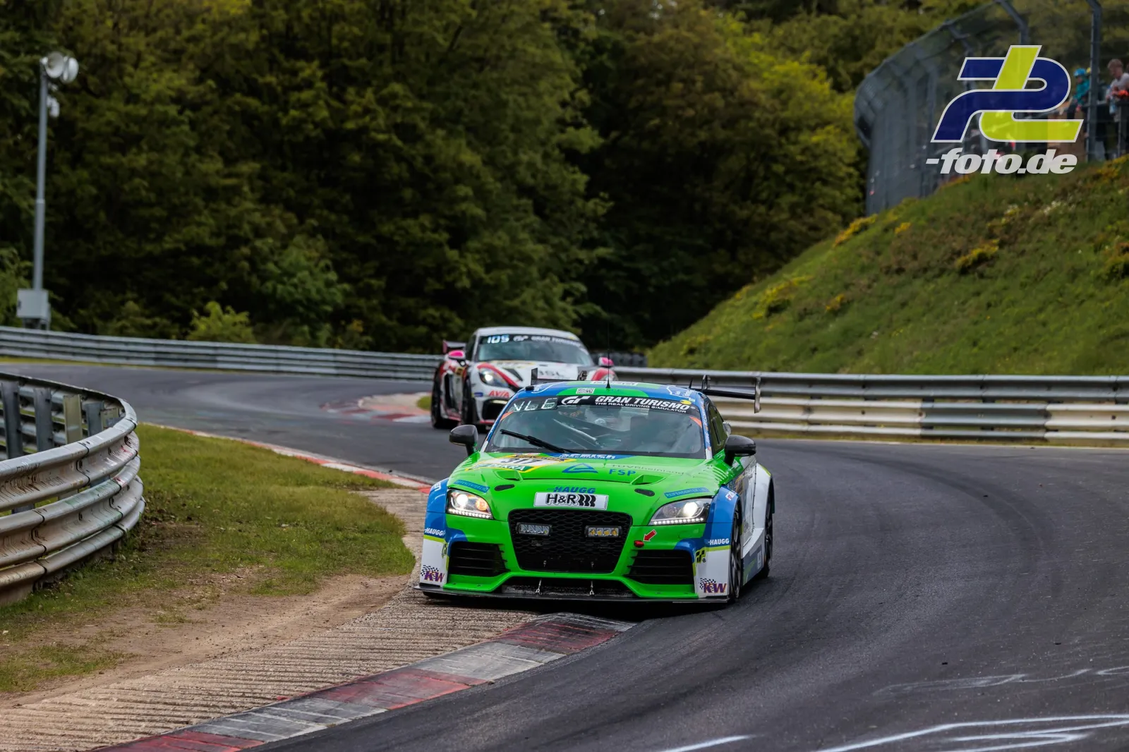 Grün-blauer Audi TTs auf der Nordschleife beim 24h-Rennen Nürburgring, verfolgt von weiterem Fahrzeug, Zuschauer im Hintergrund.