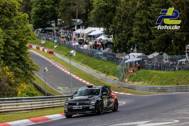 Rennfahrzeug Nr. 380 in Schwarz-Orange auf der Nürburgring-Nordschleife mit Zuschauertribünen im Hintergrund während des 24h-Rennens.