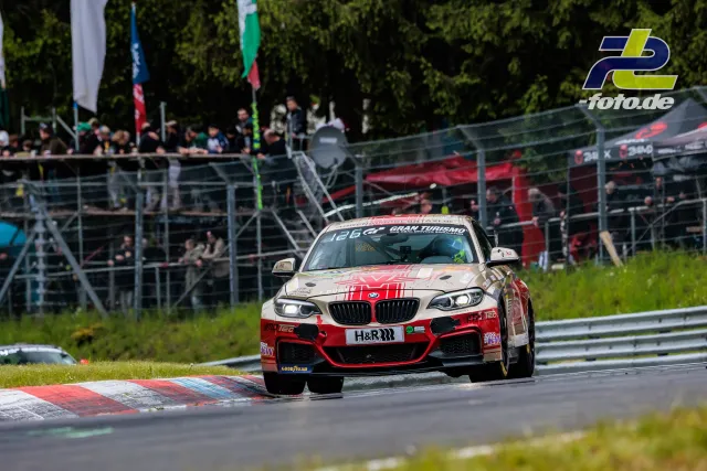 BMW M240i in rot-weißer Lackierung auf der Nordschleife des Nürburgrings, Tribüne im Hintergrund.