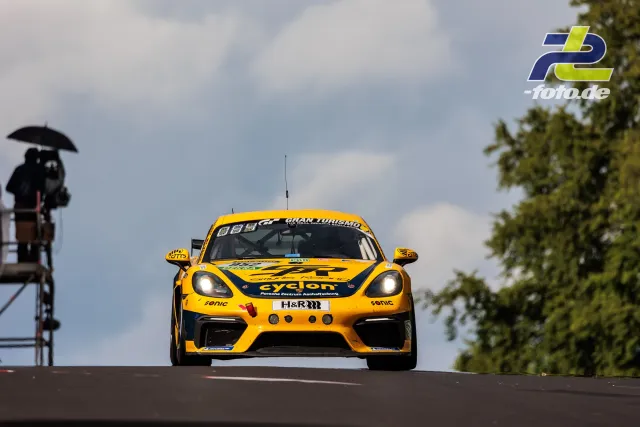 Gelber Porsche Cayman GT4 mit cyclon-Lackierung springt über eine Kuppe des Nürburgrings, im Hintergrund ein Streckenposten mit Schirm.