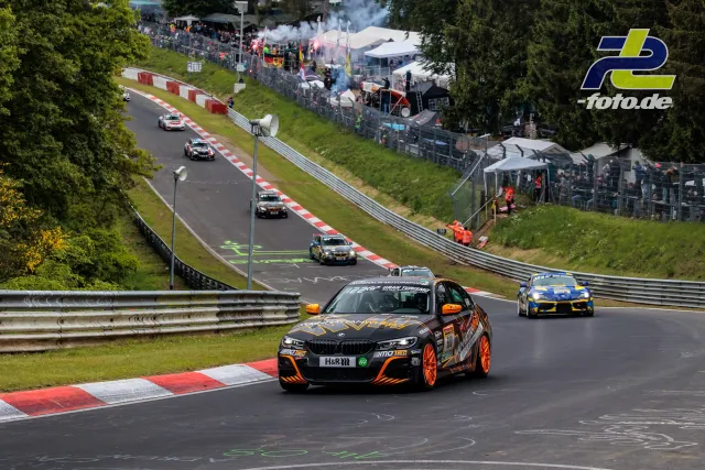 BMW mit schwarzer und oranger Lackierung führt ein Feld von Fahrzeugen durch eine Kurve auf der Nordschleife des Nürburgrings.