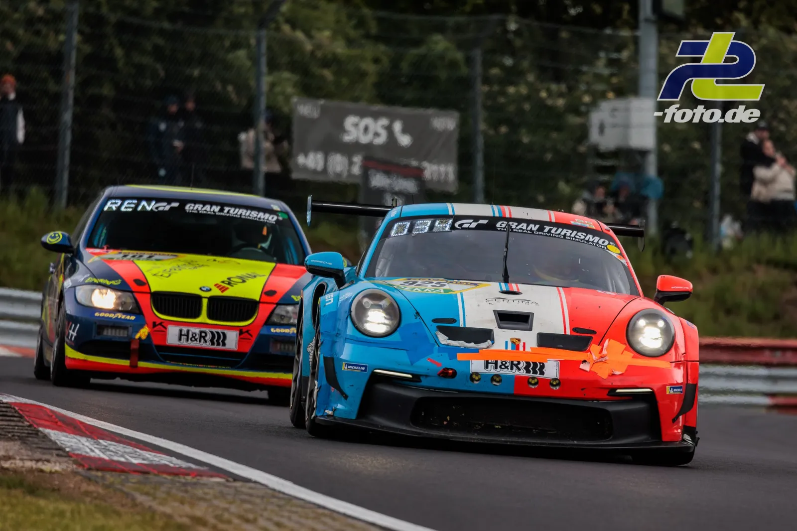 Zwei Rennwagen im Zweikampf während des 24h-Rennens auf dem Nürburgring, rechts Porsche in orange-blau-weiß, links BMW in neongelb-rot.