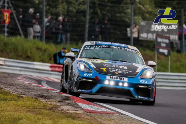 Porsche 718 Cayman GT4 #961 in blau-weißer Lackierung beim 24h-Rennen am Nürburgring mit Zuschauern im Hintergrund.