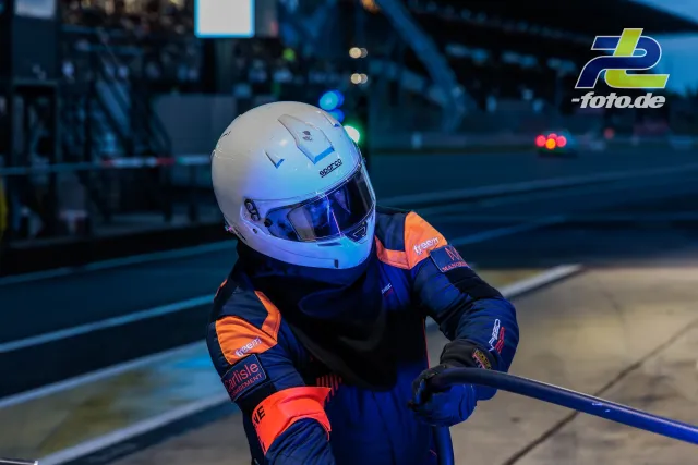 Mechaniker in blau-orangem Rennoverall mit Sparco-Helm blickt bei Dämmerung auf die Strecke, Rennwagen im unscharfen Hintergrund.