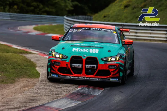 BMW M4 GT3 in türkis-roter Lackierung beim 24h-Rennen am Nürburgring in der Kurve über den Randstein fahrend.