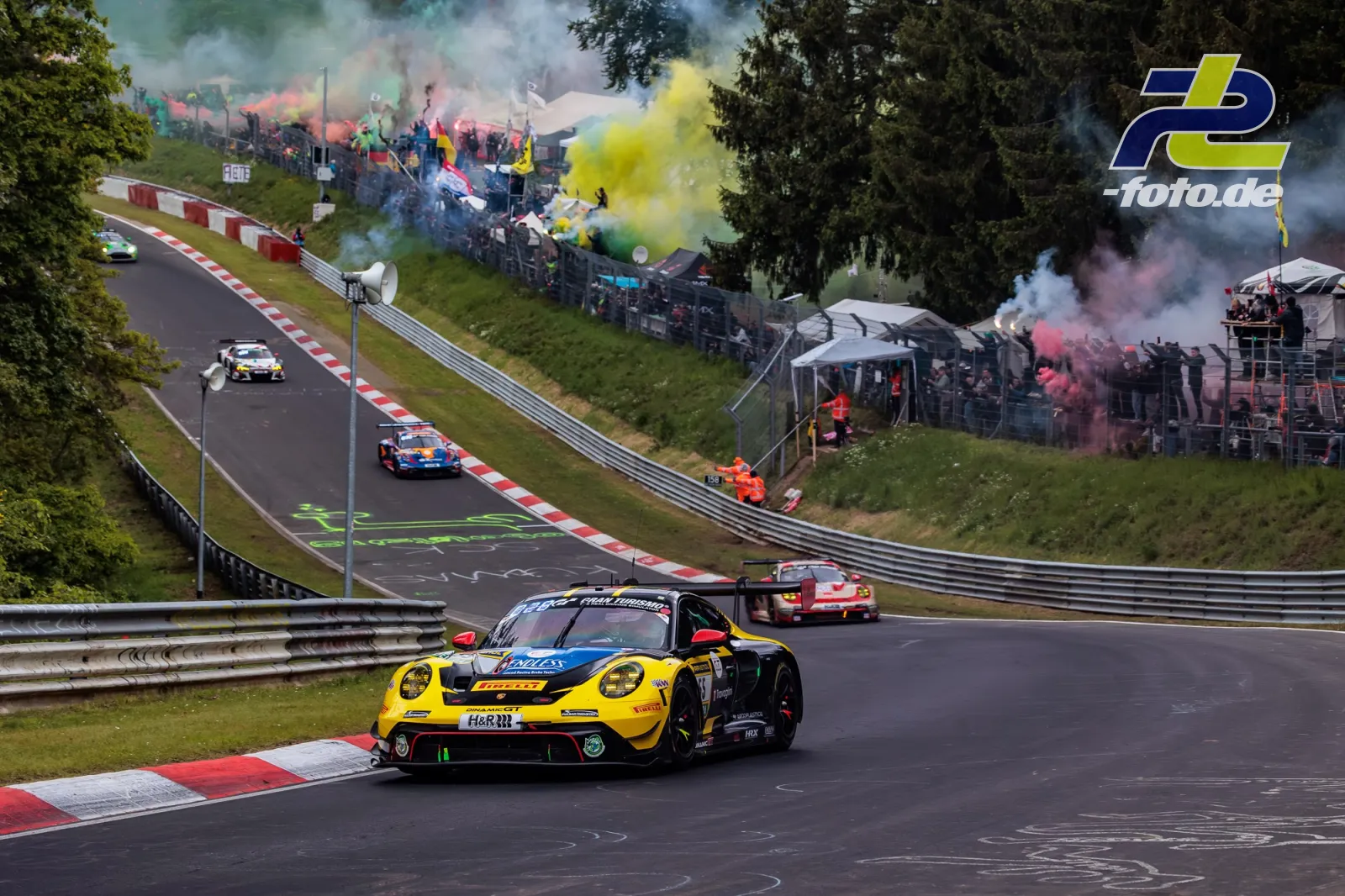 Porsche 911 GT3 R in Gelb-Schwarz auf der Nordschleife während der Formationsrunde, Zuschauer mit bunten Rauchbomben im Hintergrund