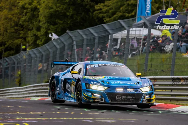 Blauer Audi R8 LMS GT3 auf der Nürburgring-Nordschleife während des 24h-Rennens, Tribüne im Hintergrund.