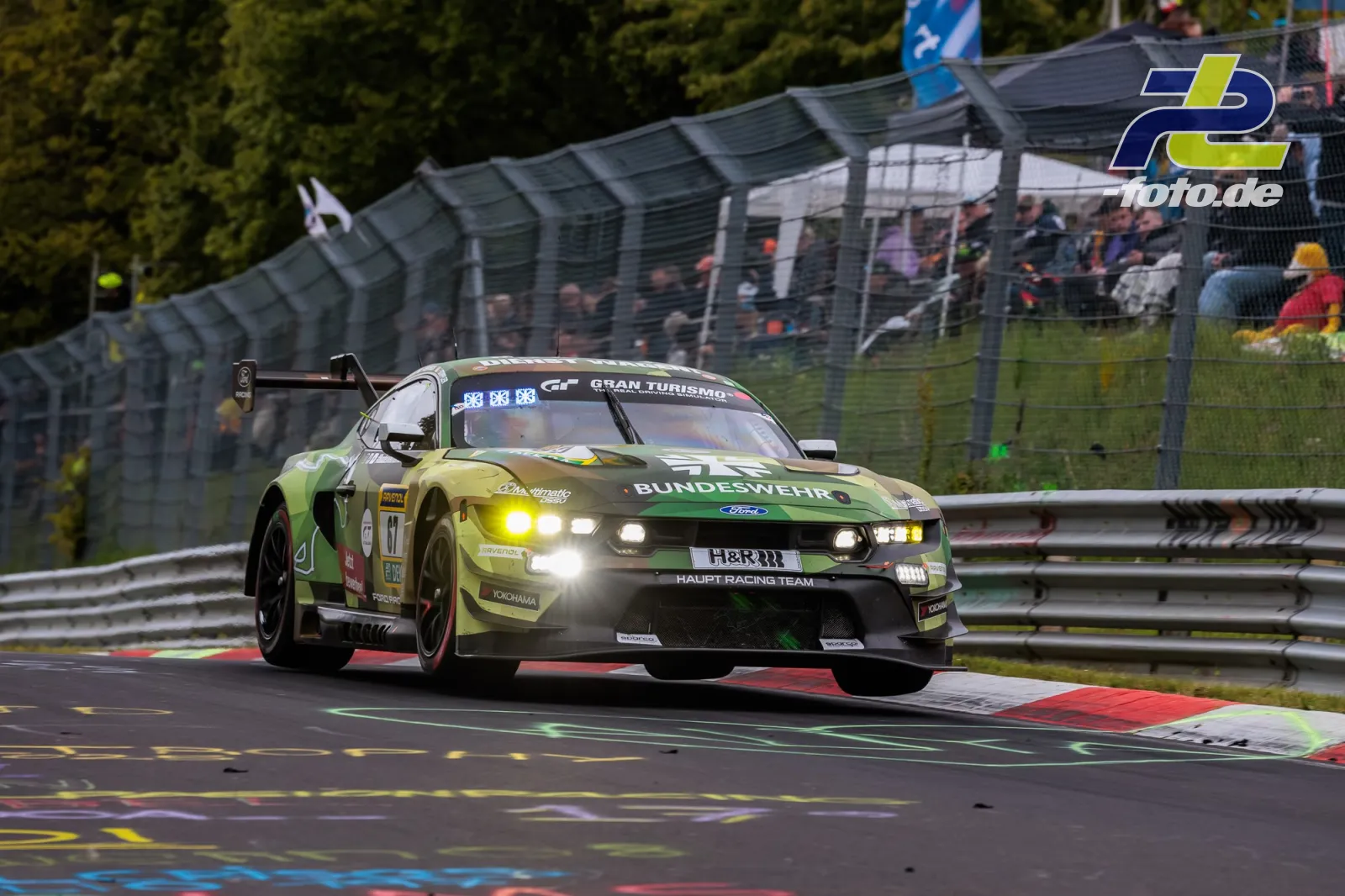 Ford Mustang GT in gelb-grüner Bundeswehr-Lackierung auf der Nordschleife des Nürburgrings mit Zuschauern im Hintergrund.