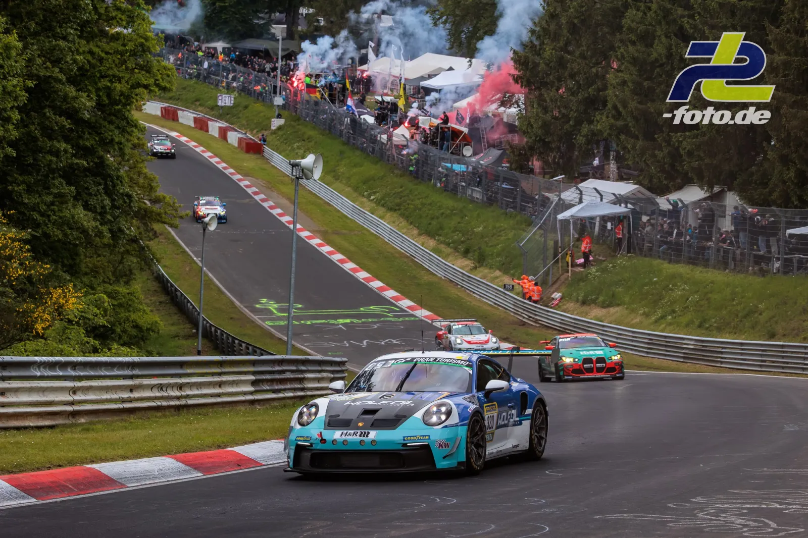 Porsche 911 GT3 in türkis-blau führt Fahrzeugfeld auf der Nürburgring-Nordschleife bei der Formationsrunde, Zuschauer mit Rauchfackeln im Hintergrund