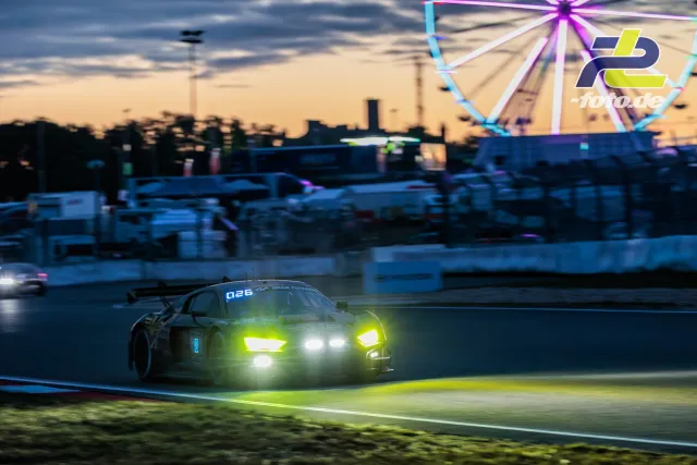 GT3-Rennwagen 8 fährt in der Dämmerung am Nürburgring, Riesenrad im Hintergrund beleuchtet, Scheinwerfer eingeschaltet.