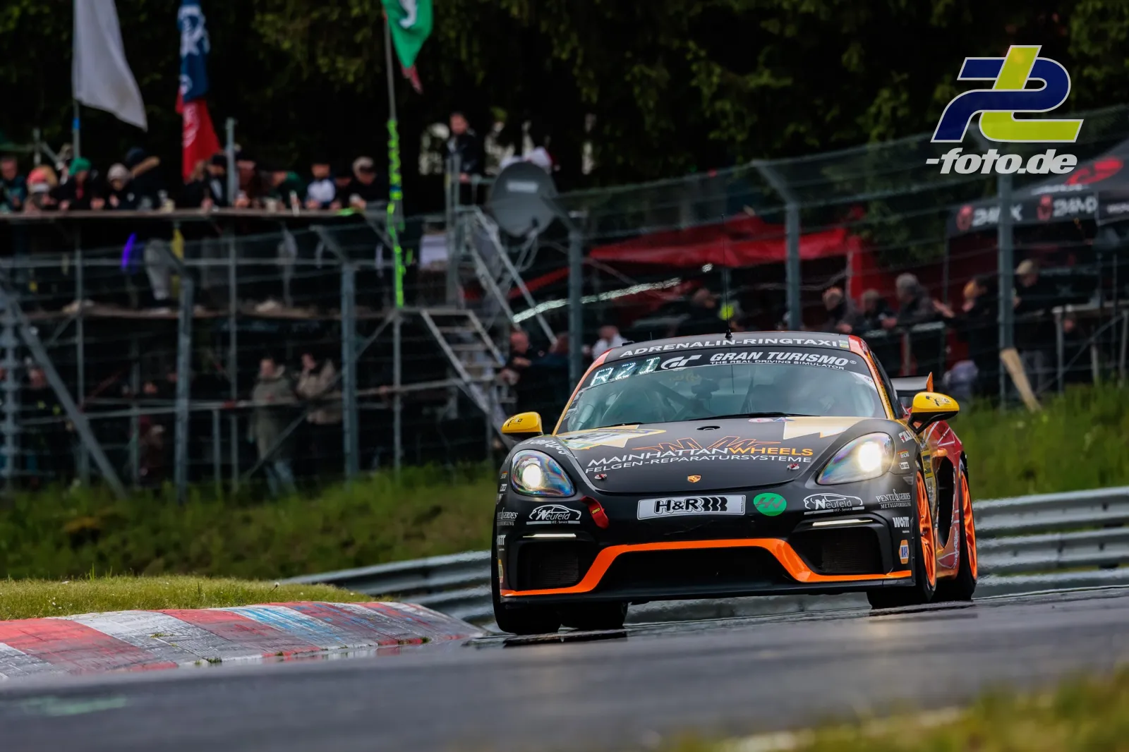 Porsche 718 Cayman GT4 Clubsport in schwarz-orange Lackierung in einer Kurve beim 24h-Rennen Nürburgring, Zuschauertribünen im Hintergrund