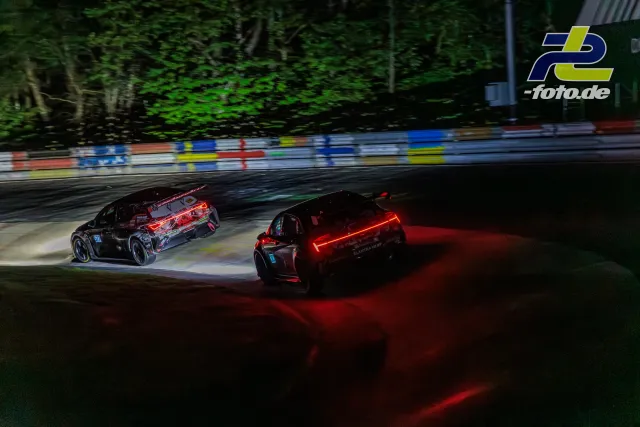 Zwei Rennwagen im Zweikampf bei Nacht auf der Nordschleife, leuchtende Bremsscheiben und Rücklichter sorgen für dramatische Lichtstimmung.