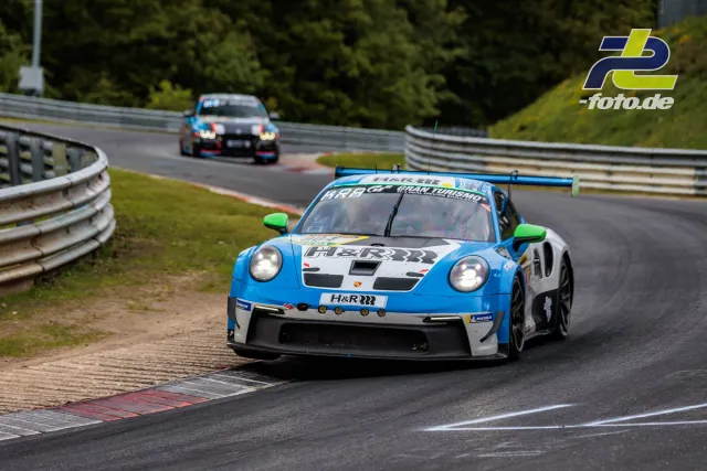 Porsche 911 GT3 R in blau-weiß-grün auf der Nordschleife, gefolgt von einem weiteren Rennwagen, Kurvenausfahrt über Randsteine.
