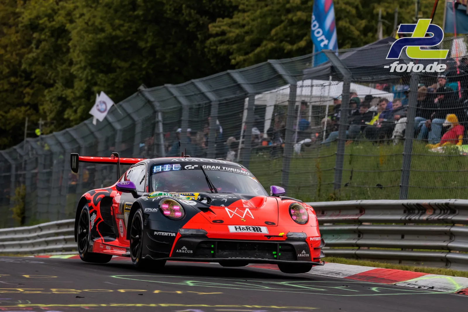 Porsche 911 GT3 R (992) Evo26 #18 von Lionspeed GP in rot-schwarz-lila während des 24h-Rennens am Nürburgring mit Zuschauern im Hintergrund