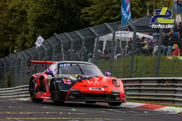 Porsche 992 GT3 R in rot-schwarzer Lackierung während des 24h-Rennens auf der Nürburgring-Nordschleife mit Zuschauern im Hintergrund.