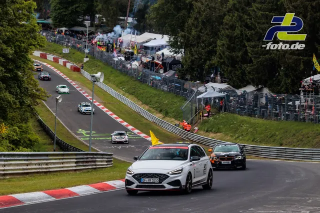 Mehrere Rennfahrzeuge auf der Nürburgring-Nordschleife, im Vordergrund weißer Hyundai und schwarzer BMW, Zuschauertribünen im Hintergrund.