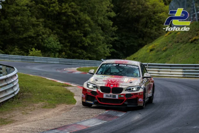 BMW M240i mit rot-weißer Lackierung fährt durch eine Kurve der Nürburgring-Nordschleife während des 24h-Rennens.