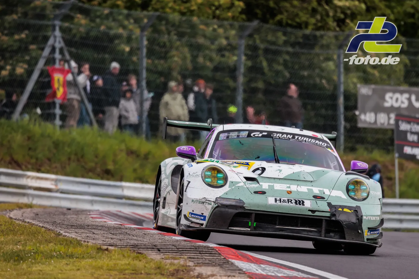 Porsche 911 GT3 R Nummer 24 in mintgrün-weißer Lackierung in einer Kurve auf dem Nürburgring, Zuschauer im Hintergrund.