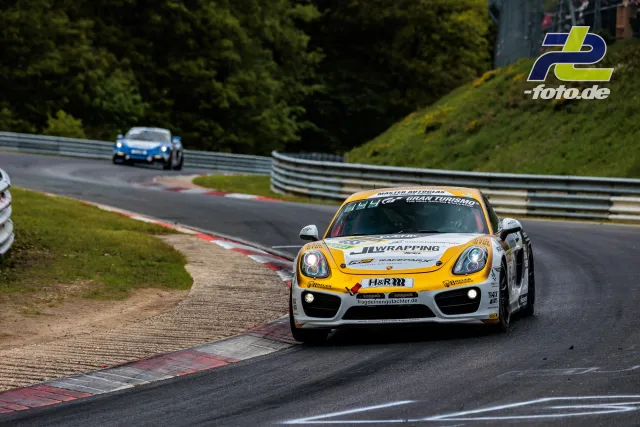 Porsche Cayman GT4 in weiß-gelb auf der Nordschleife beim 24h-Rennen, im Hintergrund ein weiterer Porsche.