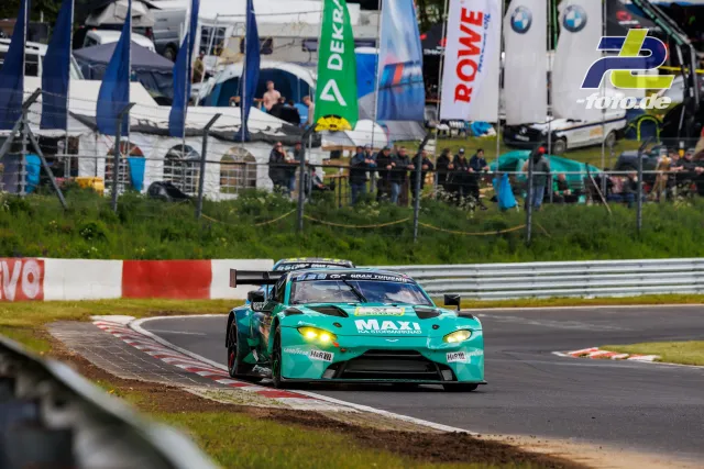Türkiser Aston Martin Vantage GT3 mit gelben Scheinwerfern auf der Nordschleife, Boxenbereich und Zuschauer im Hintergrund
