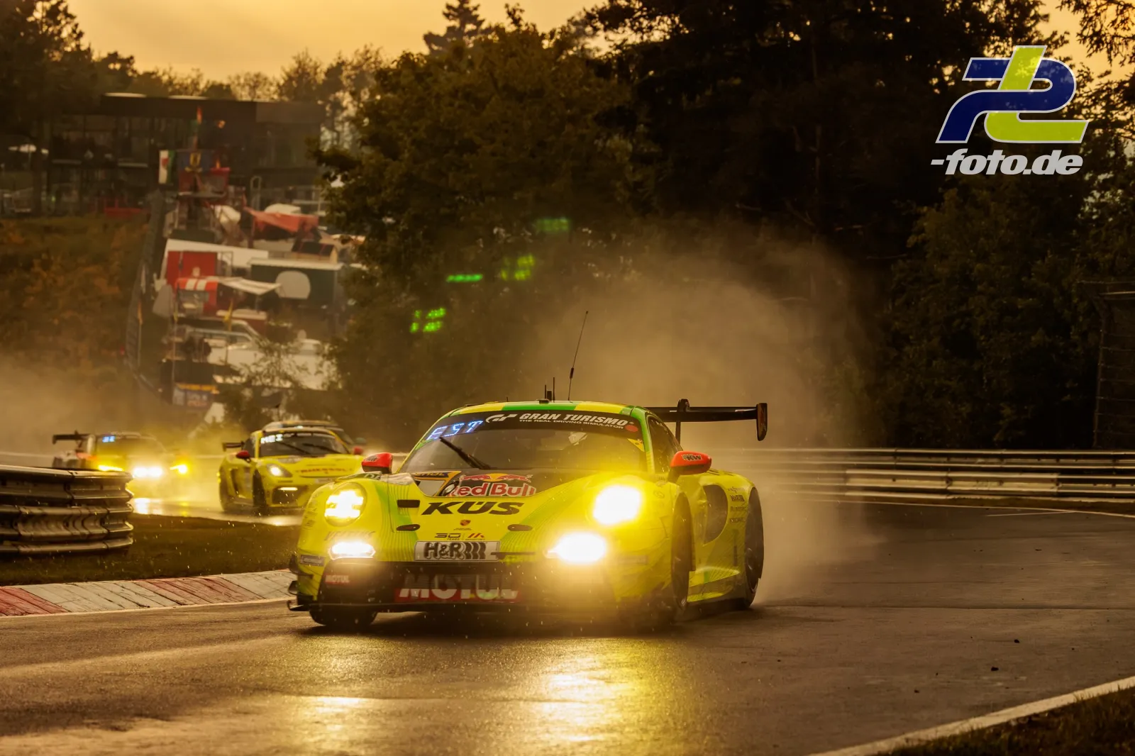 Neongelber Porsche 911 GT3 R fährt bei Dämmerung über nasse Nordschleife, gefolgt von weiteren Fahrzeugen, Gischt im Gegenlicht.