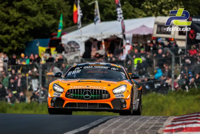 Orange-schwarzer Mercedes-AMG GT im Rennen auf dem Nürburgring, mit Zuschauern und Zelten im Hintergrund.