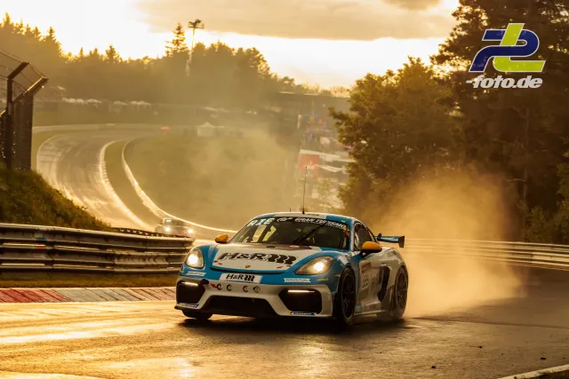 Porsche Cayman GT4 in Kurve auf nasser Nordschleife bei Gegenlicht und Dunst, Sonnenuntergang über bewaldeter Strecke.