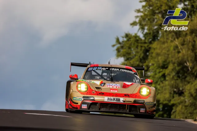Porsche 911 GT3 R in gold-roter Lackierung über eine Kuppe am Nürburgring, eingeschaltete Scheinwerfer, Bäume im Hintergrund.