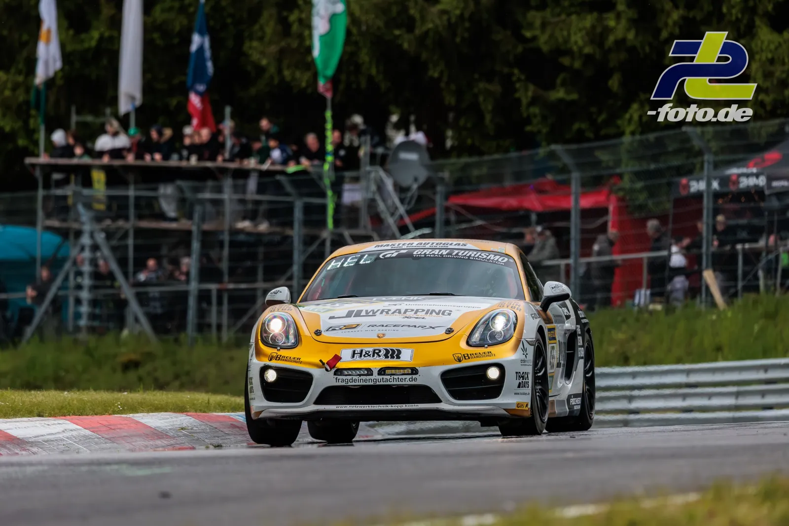 Porsche Cayman in orange-weißer Lackierung auf der Nordschleife während des 24h-Rennens, Zuschauertribüne im Hintergrund.