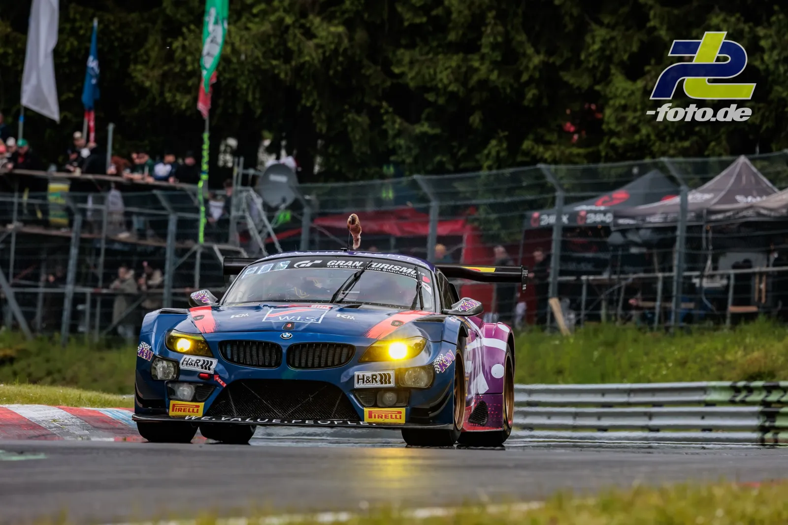 BMW Z4 GT3 mit markanter blau-pinker Lackierung während des 24h-Rennens auf dem Nürburgring mit Boxengasse im Hintergrund.