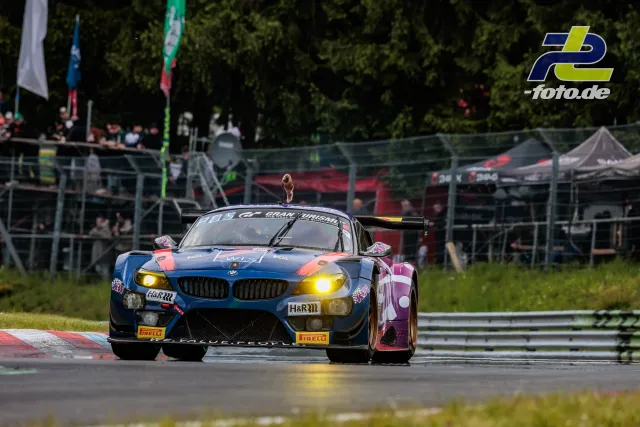BMW Z4 GT3 mit markanter blau-pinker Lackierung während des 24h-Rennens auf dem Nürburgring mit Boxengasse im Hintergrund.