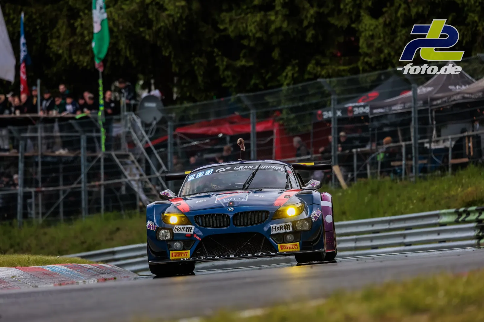 BMW E89 Z4 in blau-pink auf der Nordschleife beim 24h-Rennen, Zuschauer im Hintergrund, gelbe Scheinwerfer leuchten.