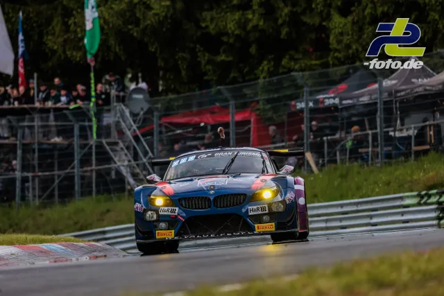 BMW Z4 GT3 in blau-roter Lackierung auf der Nordschleife mit Tribüne und Zuschauern im Hintergrund während des 24h-Rennens