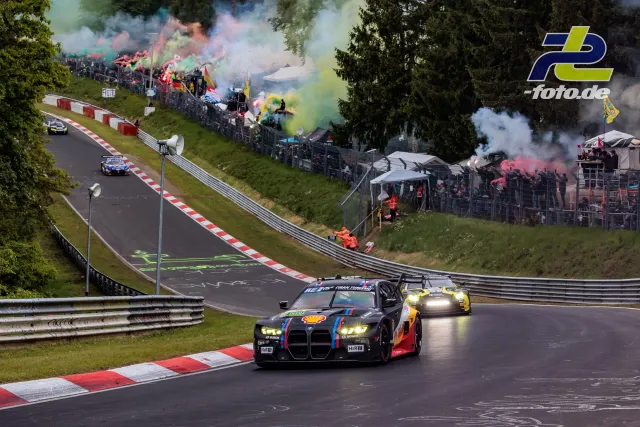 BMW M4 GT3 mit markanter Nierengrill-Lackierung führt das Feld auf der Nordschleife durch Kurvenpassage mit bunten Rauchfackeln der Fans.